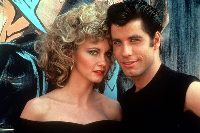 Two people pose closely together, dressed in retro style, against a colorful mural background. One has curly hair; the other has slicked-back hair.