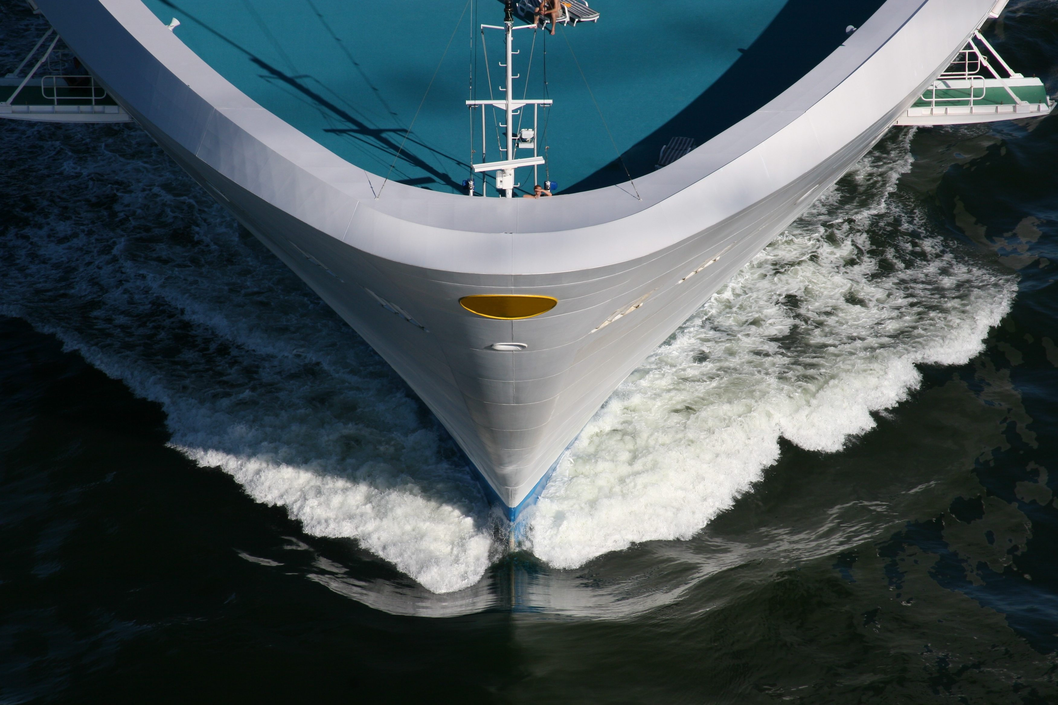 A ship cuts through water, creating waves, with a blue deck visible, in a maritime setting.