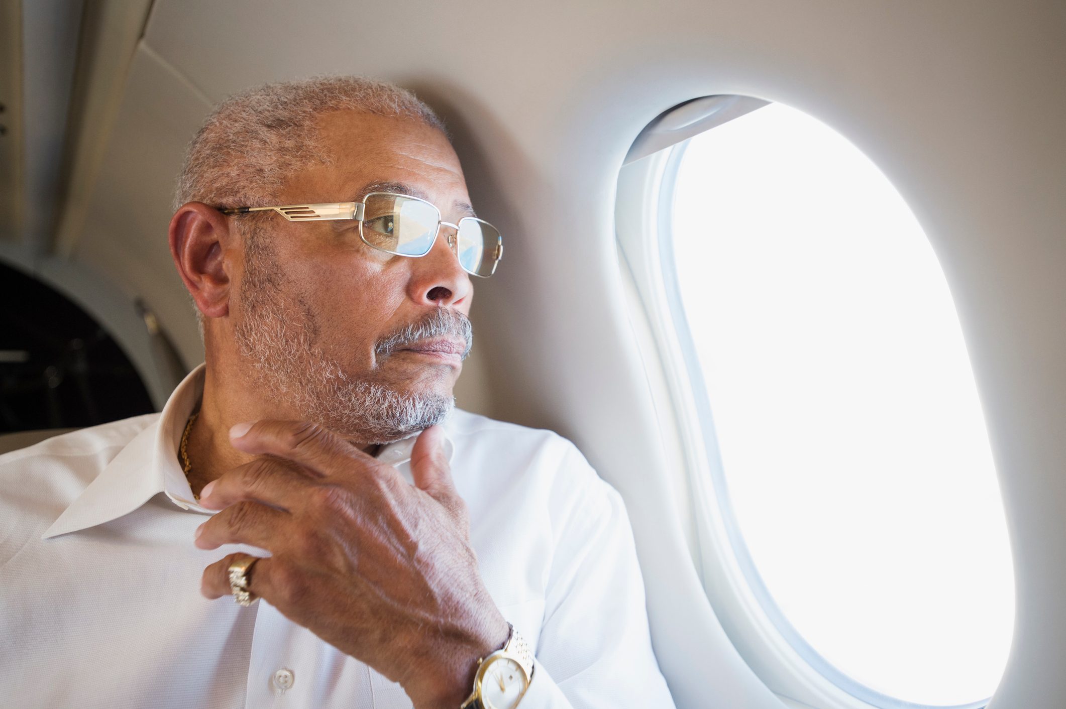 A person looks out an airplane window, wearing glasses and a white shirt, with a thoughtful expression.