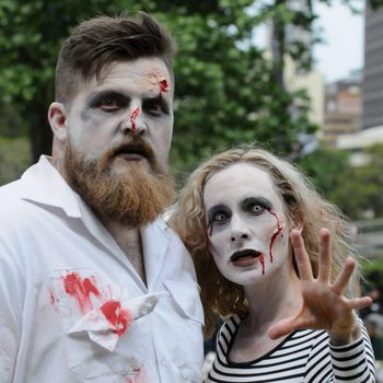 Two people are dressed as zombies, standing outdoors with makeup and fake blood, against a backdrop of trees and buildings.