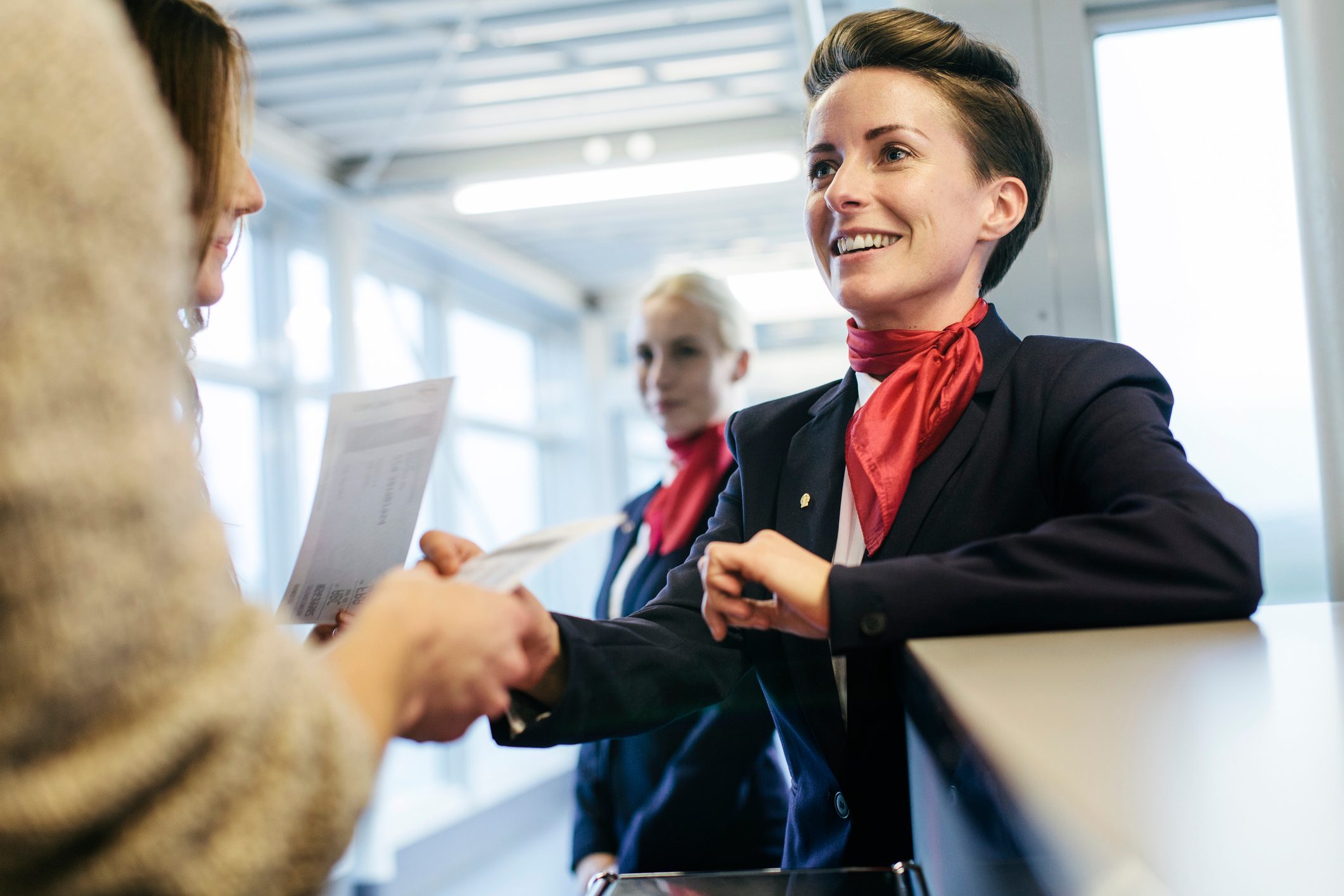A smiling airline attendant assists a passenger at a check-in counter, with a colleague in the background at an airport.