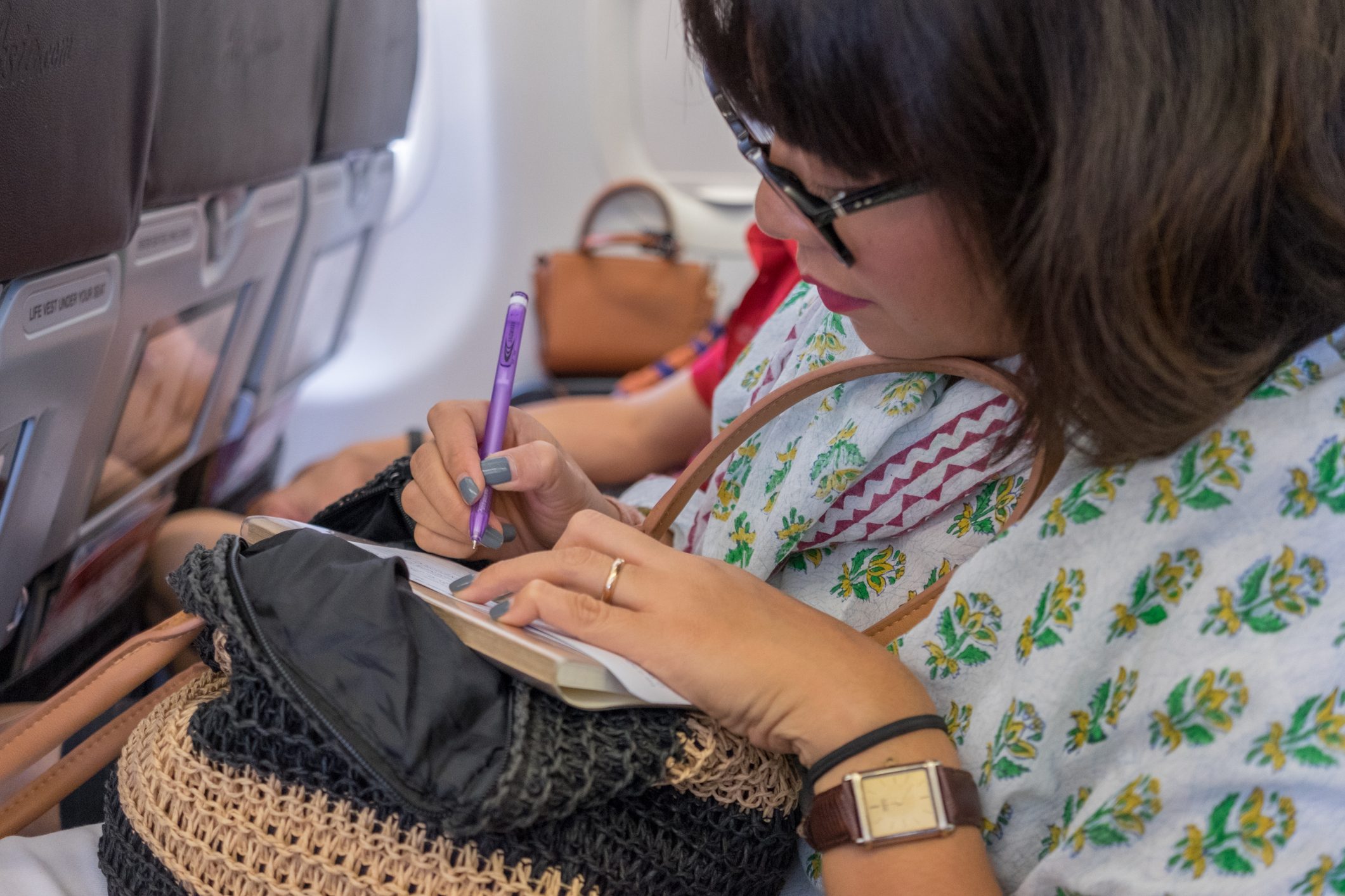 Person writes with a purple pen in a small notebook, seated on an airplane with other passengers in the background.