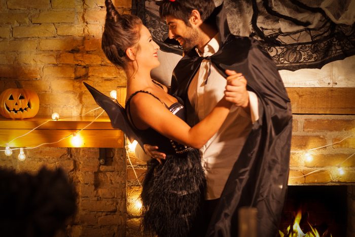 Couple dances in Halloween costumes by a brick fireplace with string lights and a carved pumpkin on the mantel.