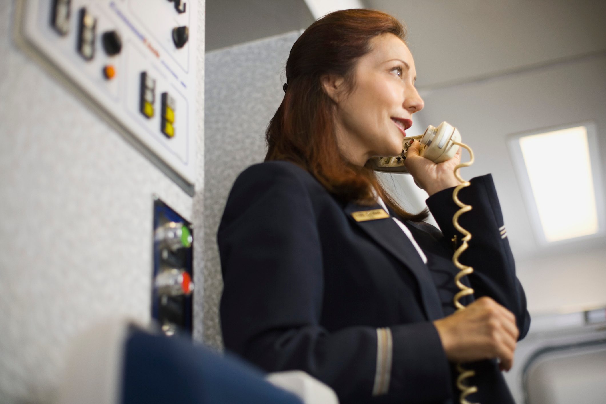 Flight attendant speaks on a handset inside an airplane, surrounded by control panels and cabin interior.
