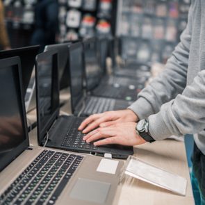 Laptops display in a row; person's hands adjust one. Store environment with blurred shelves in background.