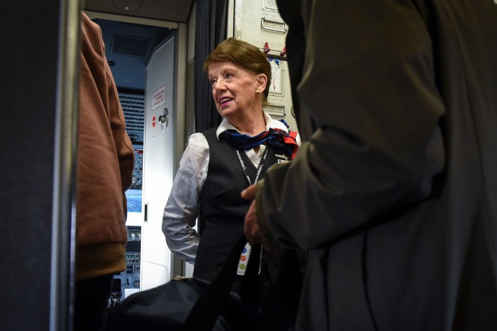 Flight attendant converses with passengers near the cockpit doorway, wearing a uniform with a scarf, inside an airplane cabin.
