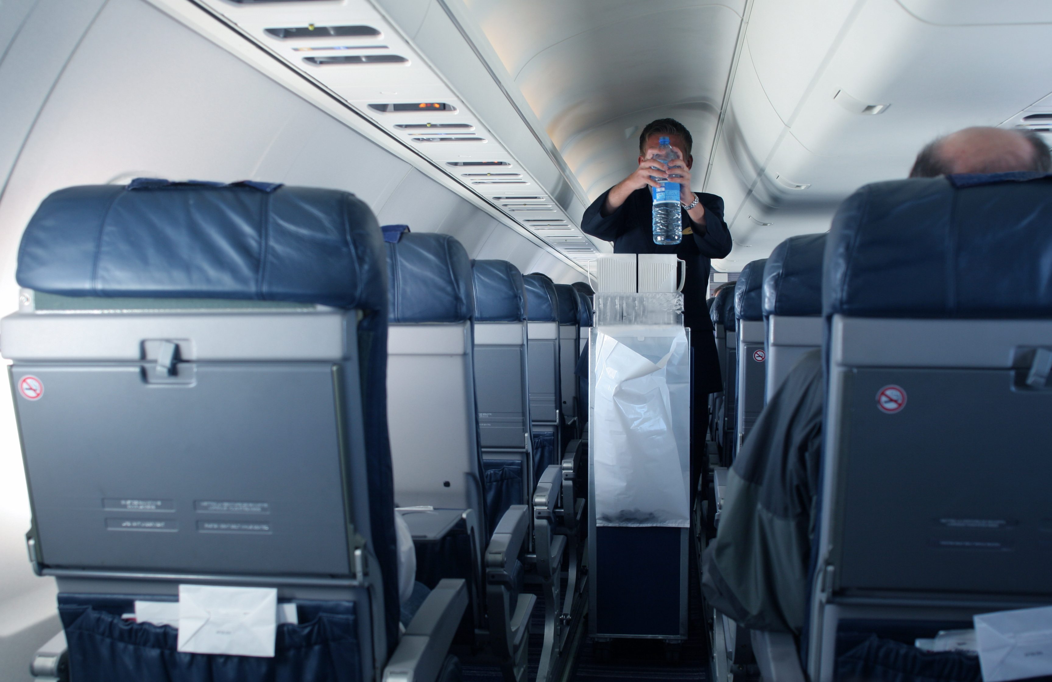 Flight attendant holds water bottle in aircraft aisle, surrounded by seated passengers and tray tables with no-smoking signs visible.