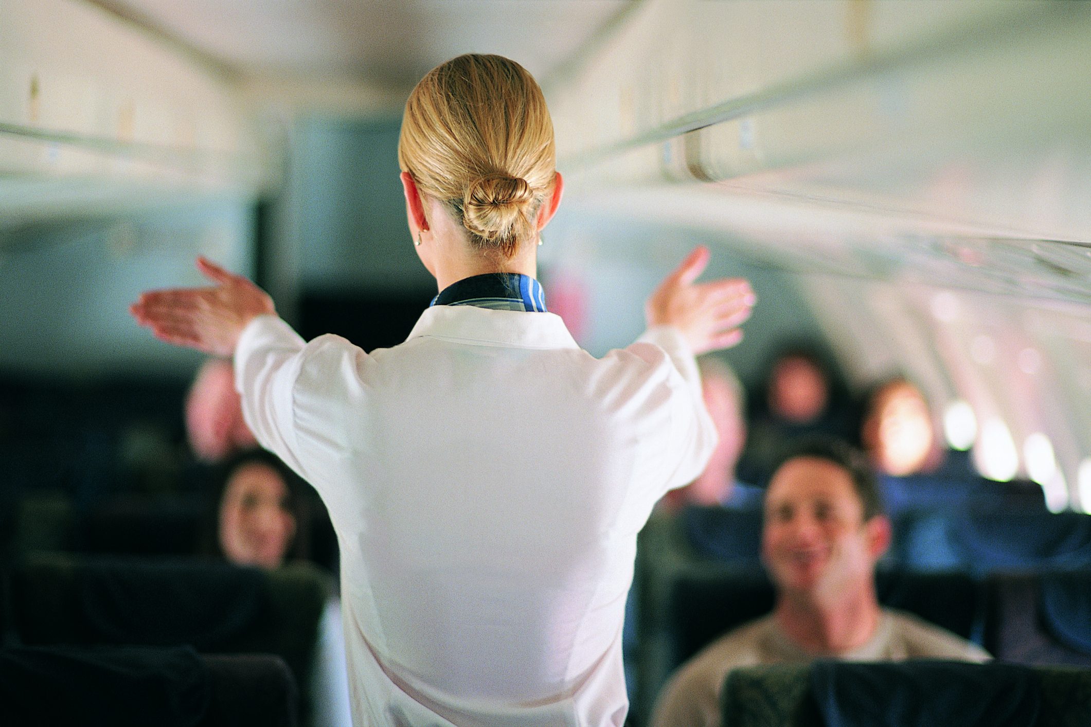 Flight attendant gestures during safety demonstration, with passengers seated attentively inside an airplane cabin.