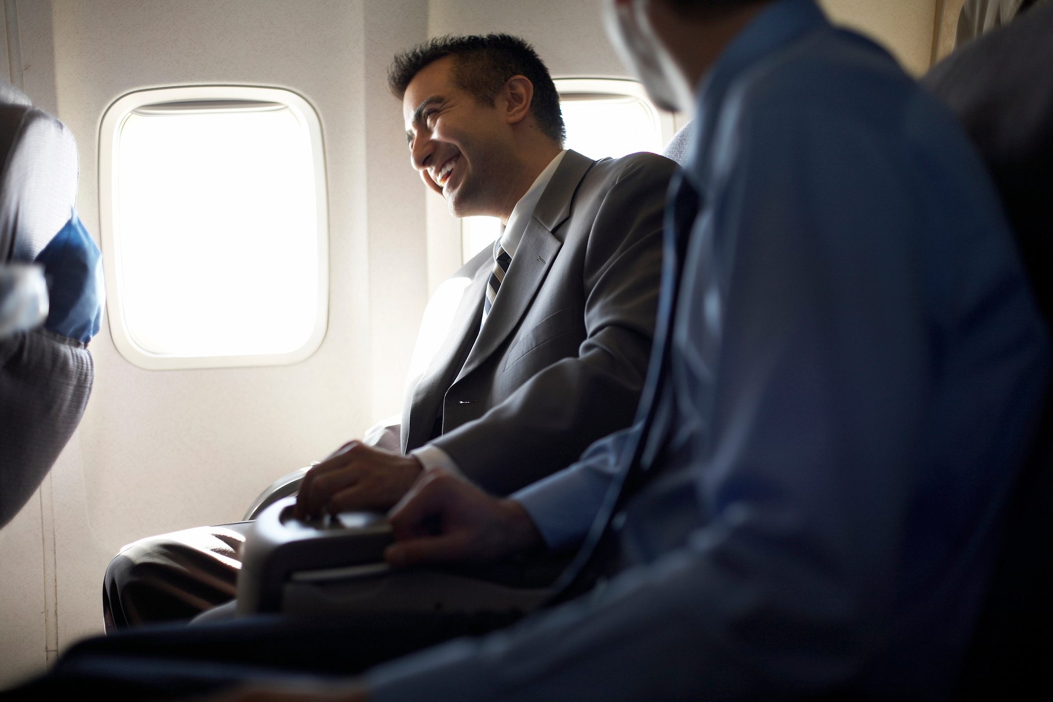 Man smiling and sitting on an airplane, talking to a nearby person, with bright light streaming through the window.