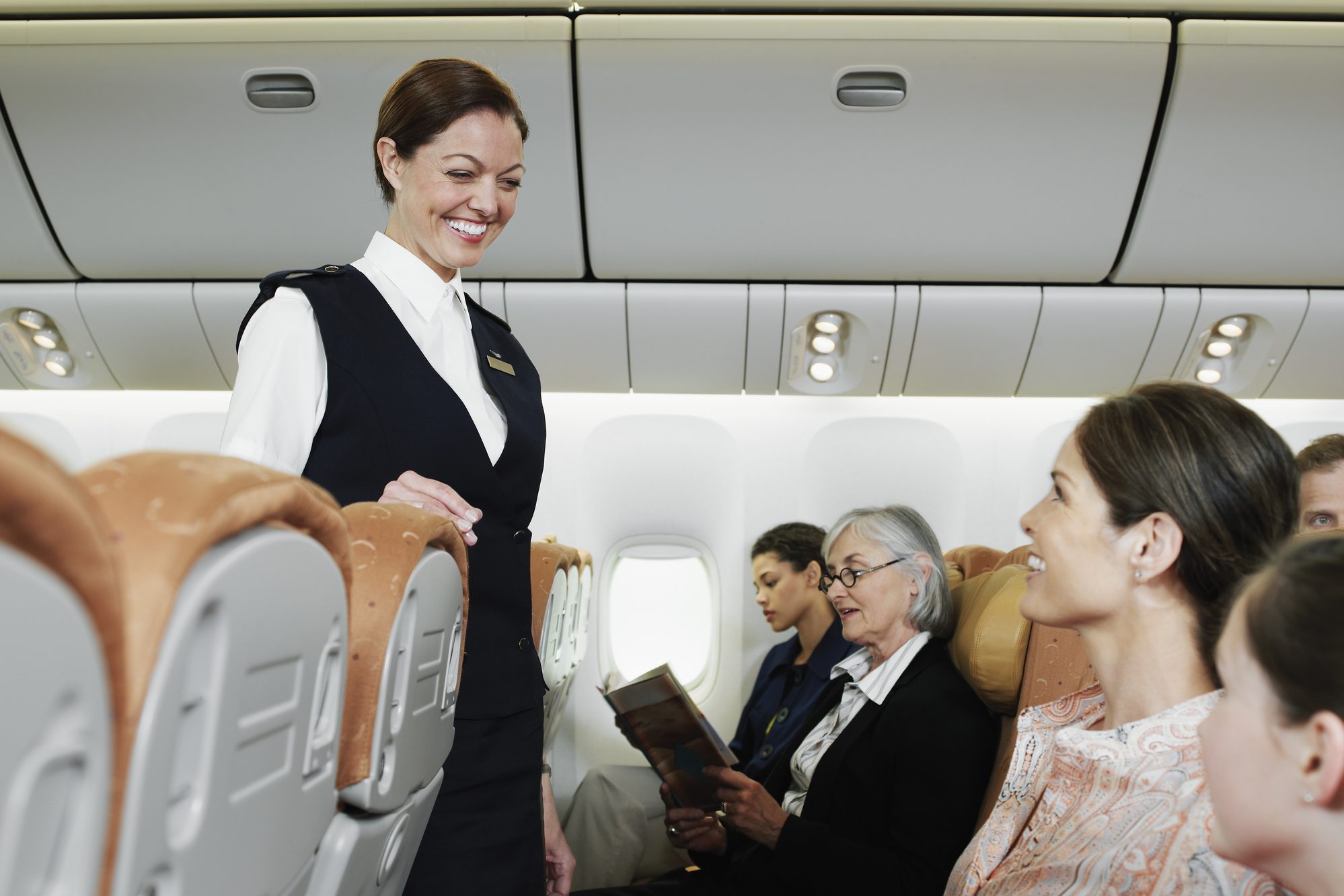 Flight attendant smiling, interacting with passengers seated in an airplane cabin. Two women are reading, and a child is looking forward attentively.