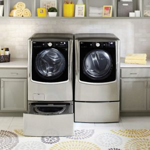 Front-loading washers stand side-by-side; one drawer is open. Set in a neatly organized laundry room with shelves, cabinets, and decorative items.