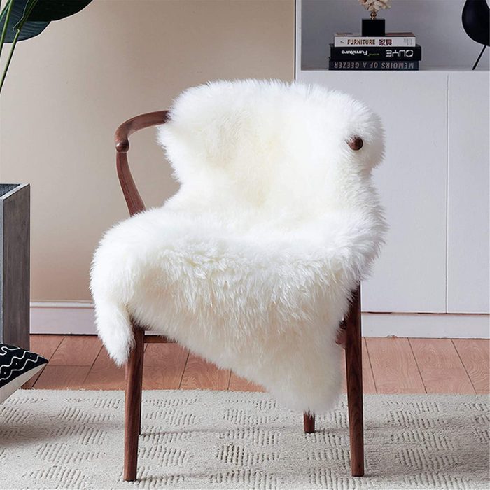 A wooden chair covered with a fluffy white fur lies beside a modern shelf containing stacked books in a cozy room.