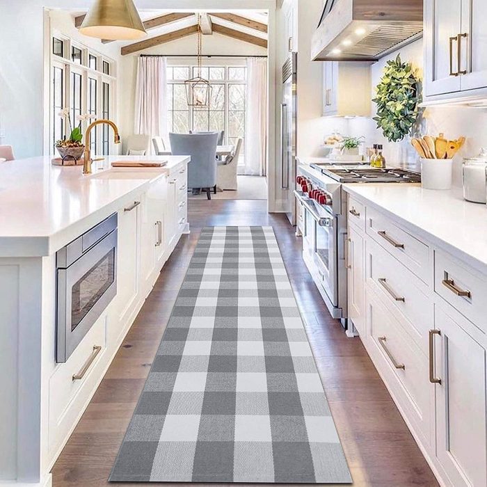 Runner rug lies on wooden floor in a bright kitchen, featuring white cabinets and appliances, leading to a dining area with large windows.