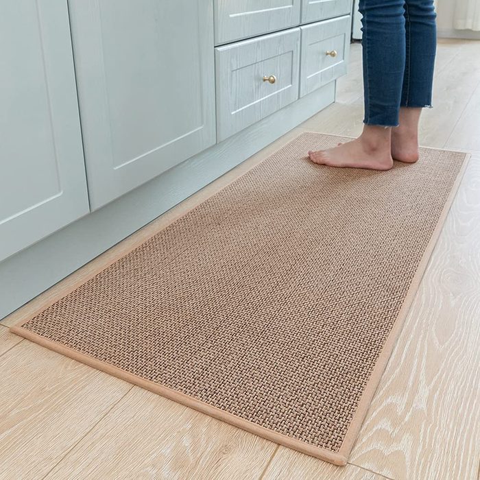 A person stands barefoot on a woven mat in a bright kitchen with light wood flooring and pale cabinetry.