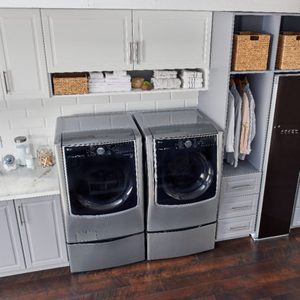 Two silver front-loading washers sit side by side. Next to them, shelves hold towels, baskets, and hanging clothes in a tidy laundry room.