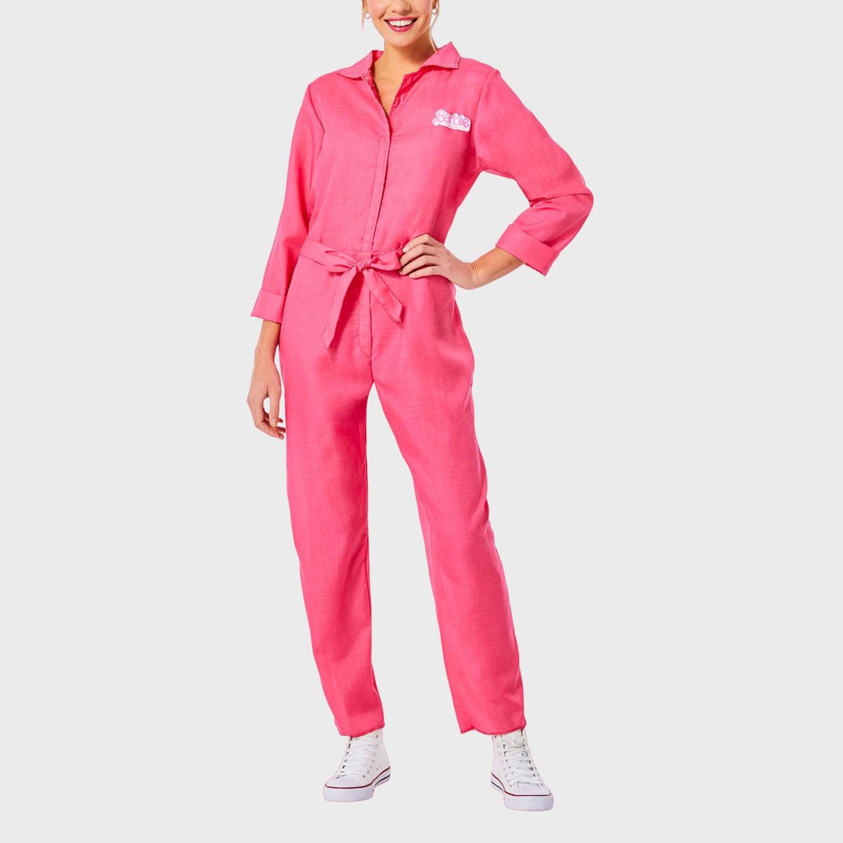 Person stands confidently wearing a pink jumpsuit with "Barbie" text, white sneakers, against a plain white background.