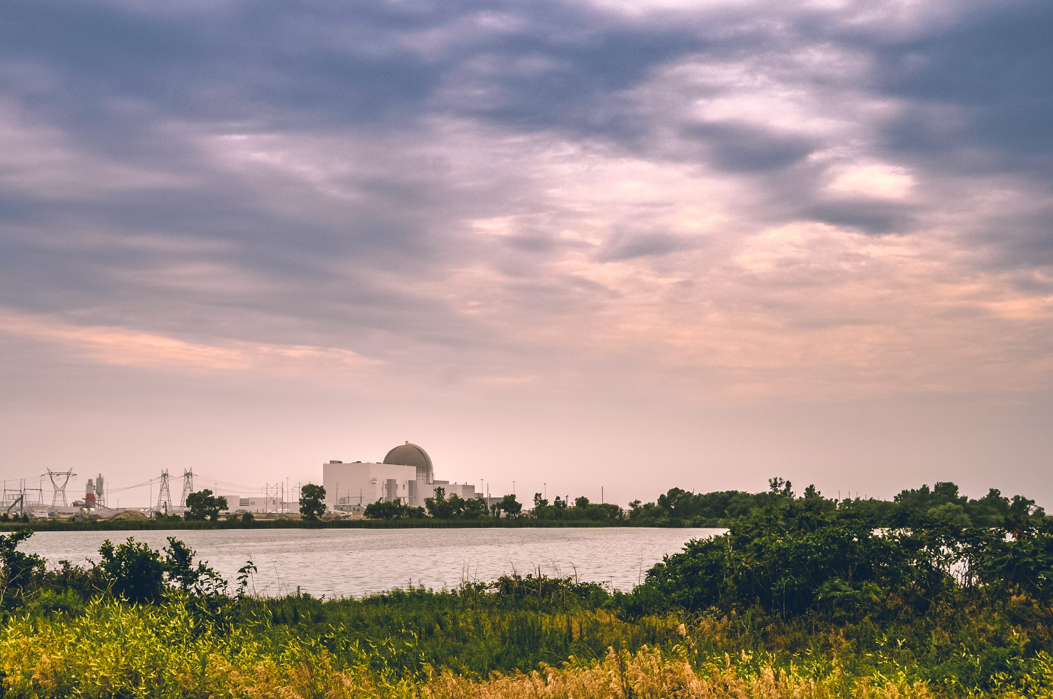 A power plant stands quietly, surrounded by water, greenery, and an overcast sky, creating a serene industrial landscape.