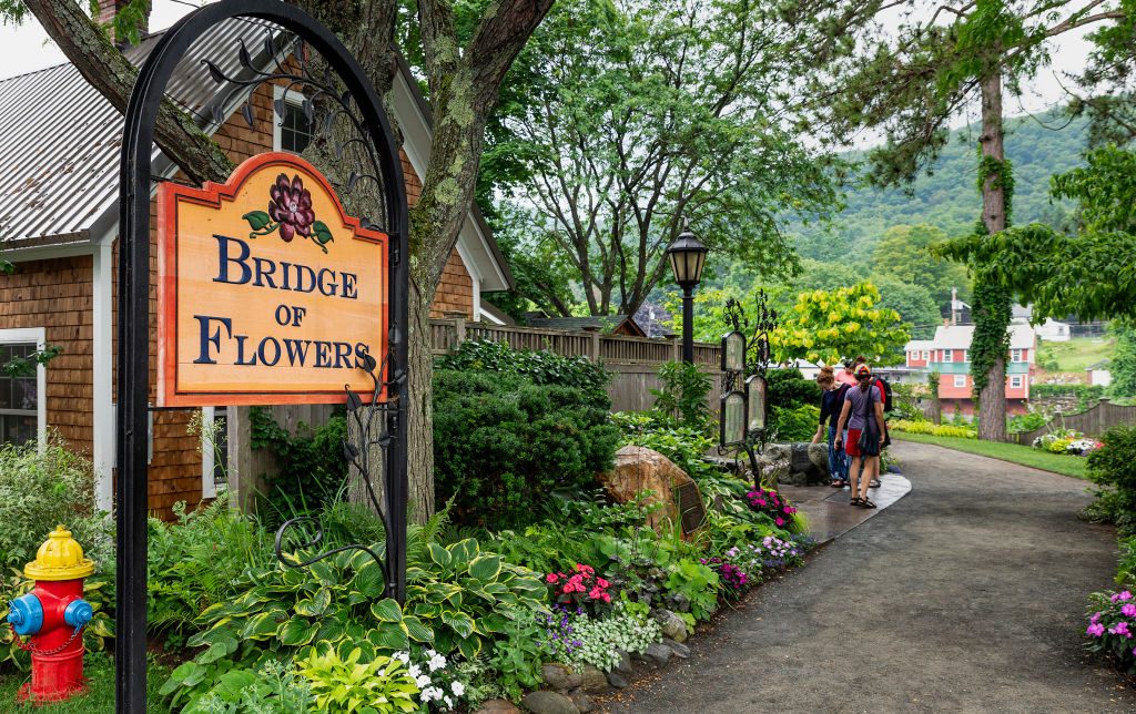 Sign reads "Bridge of Flowers" beside a pathway with people walking, surrounded by lush gardens and trees, near a rustic house.