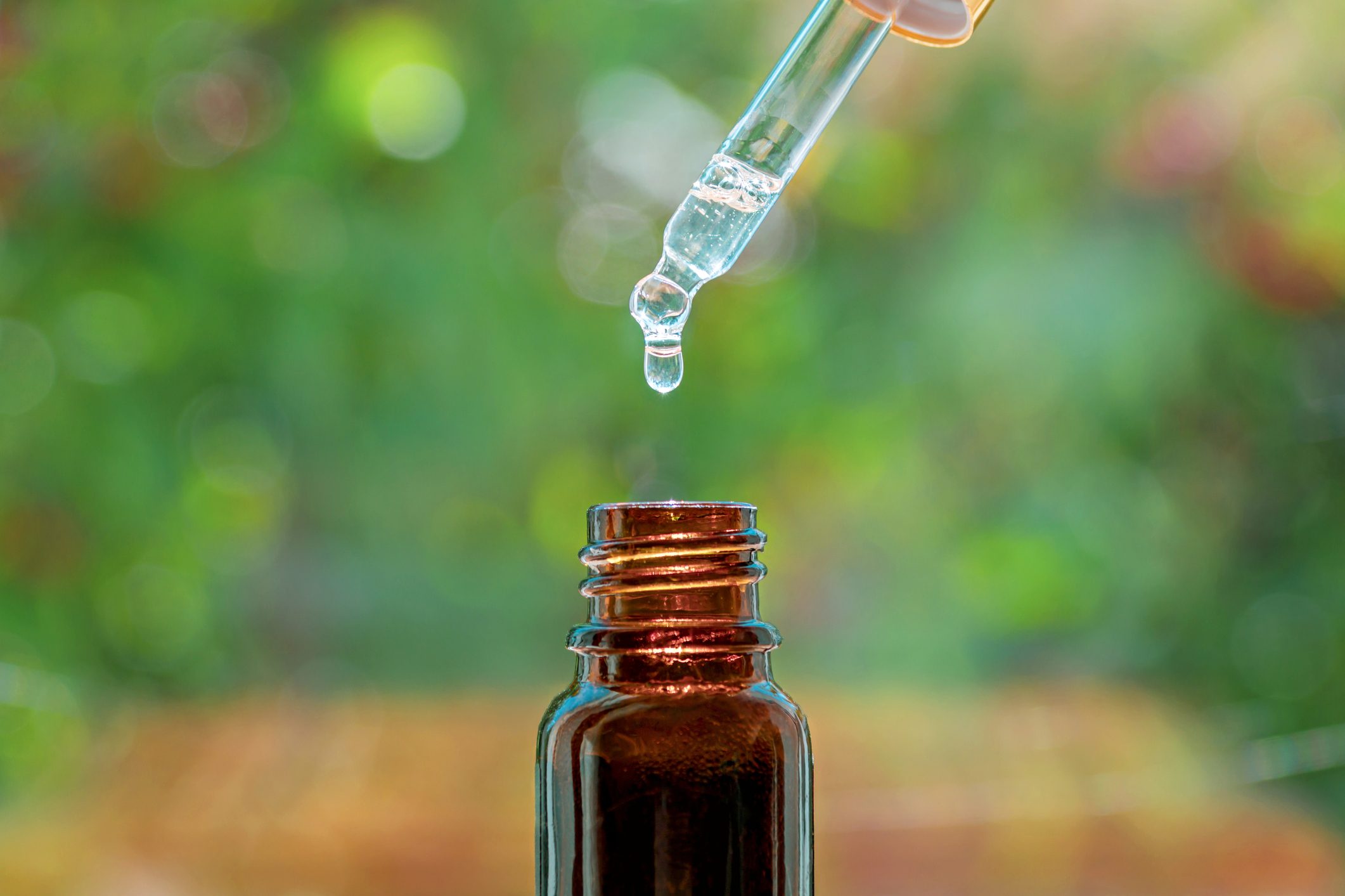 Glass dropper releasing liquid into a small, brown bottle with a blurred green and colorful background.