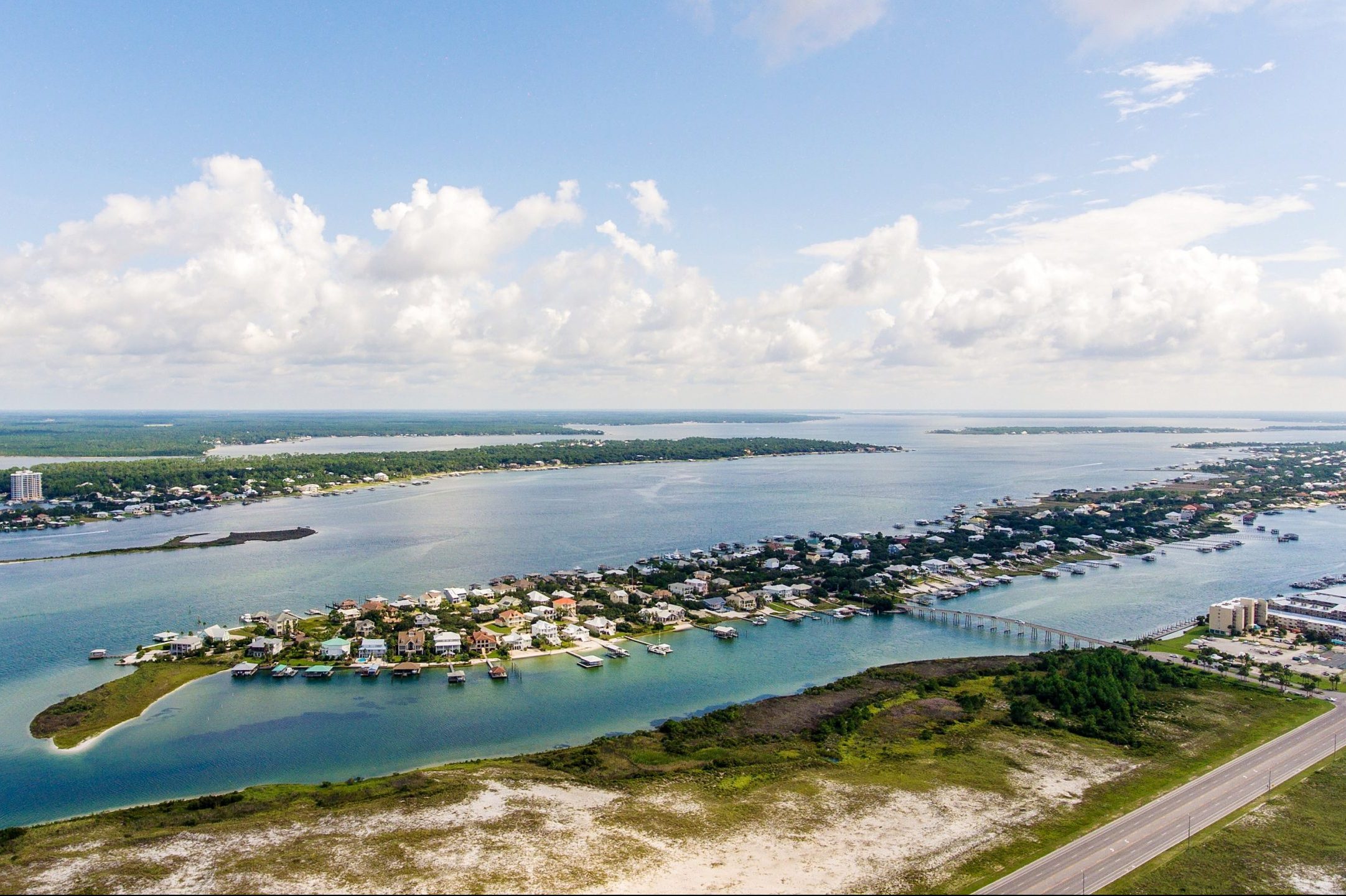 Aerial view of a coastal town, nestled along a winding river, with houses lining the shoreline and green land beyond under a partly cloudy sky.