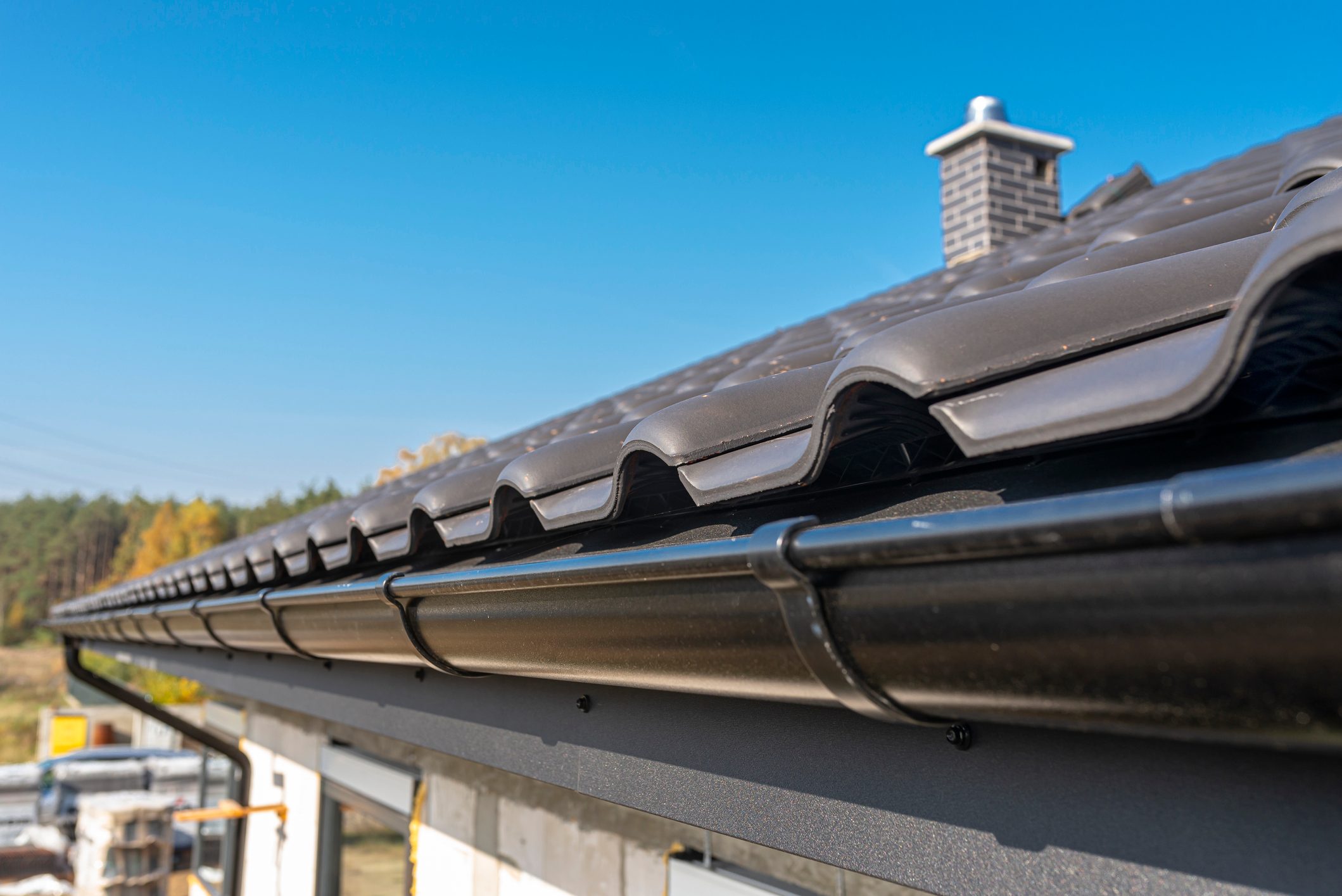 Black roof tiles rest on a sloped roof, with a metal gutter beneath, houses are nearby, and trees are visible under a clear blue sky.