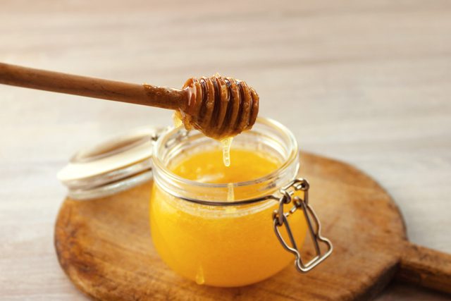 A honey dipper drips honey into an open glass jar placed on a wooden cutting board in a kitchen.