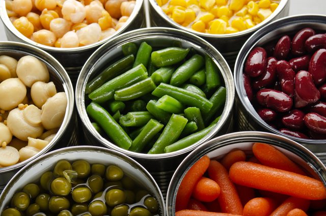 Open metal cans display colorful vegetables like peas, carrots, beans, and corn arranged in a vibrant, appetizing presentation.
