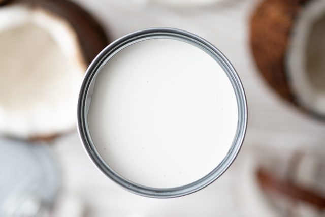 A can filled with white coconut milk sits on a table, surrounded by blurred coconut shells.