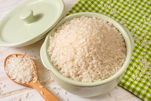 A bowl of uncooked rice sits on a green checkered cloth, accompanied by a wooden spoon with rice grains.
