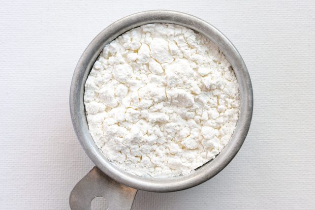 Metal measuring cup holds white, powdery substance; placed on a textured, light surface.