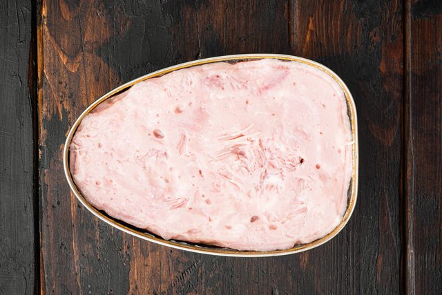 Oval can contains pink luncheon meat, nestled on dark, textured wooden surface.