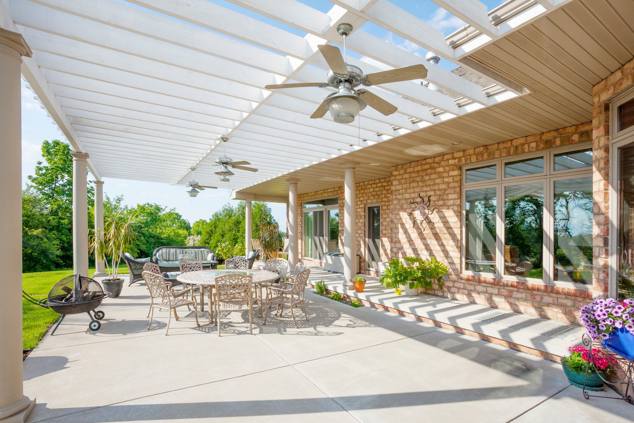 Patio features round table with chairs under pergola, surrounded by greenery, ceiling fans, potted plants, and a fire pit on concrete surface.