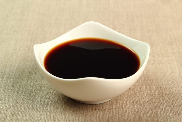 A white bowl holds dark soy sauce, resting on a beige fabric surface.