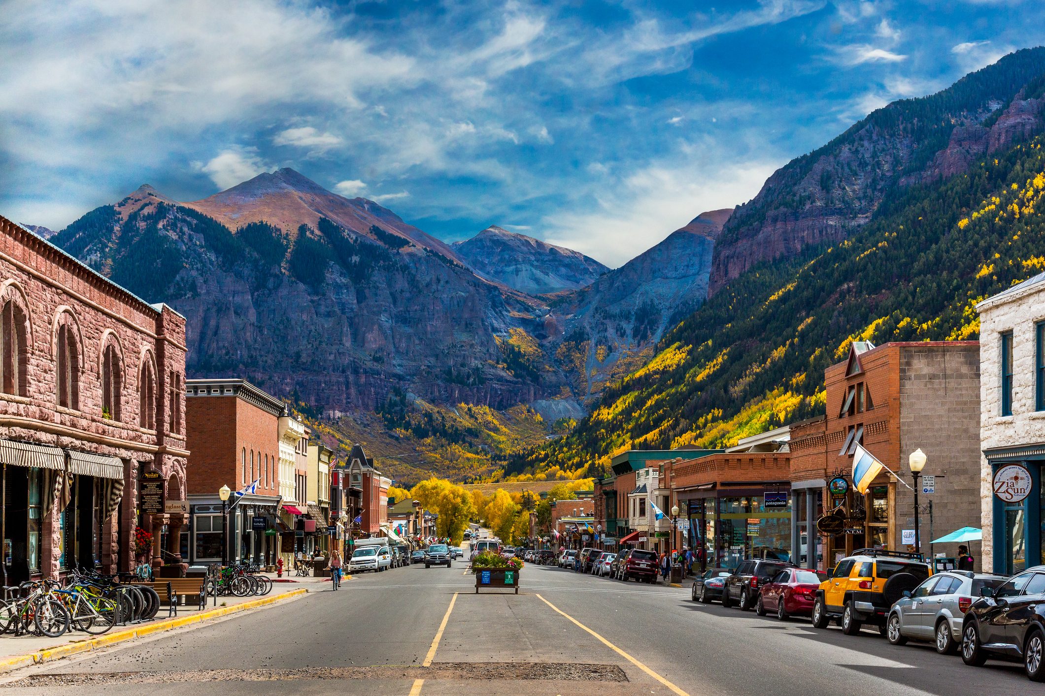 Street lined with colorful shops and parked cars, leading to mountains with yellow foliage under a partly cloudy sky.