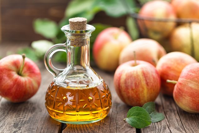Glass bottle filled with amber liquid, cork sealed, surrounded by red apples on a wooden surface with green leaves nearby.