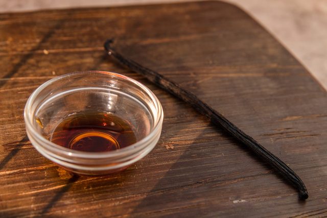 Glass bowl holds dark liquid; a vanilla bean rests beside it on a wooden surface.