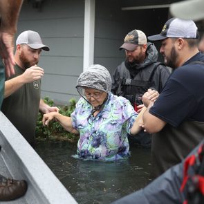 A woman gets assisted by several men out of floodwaters near a building, all are standing in waist-deep water.