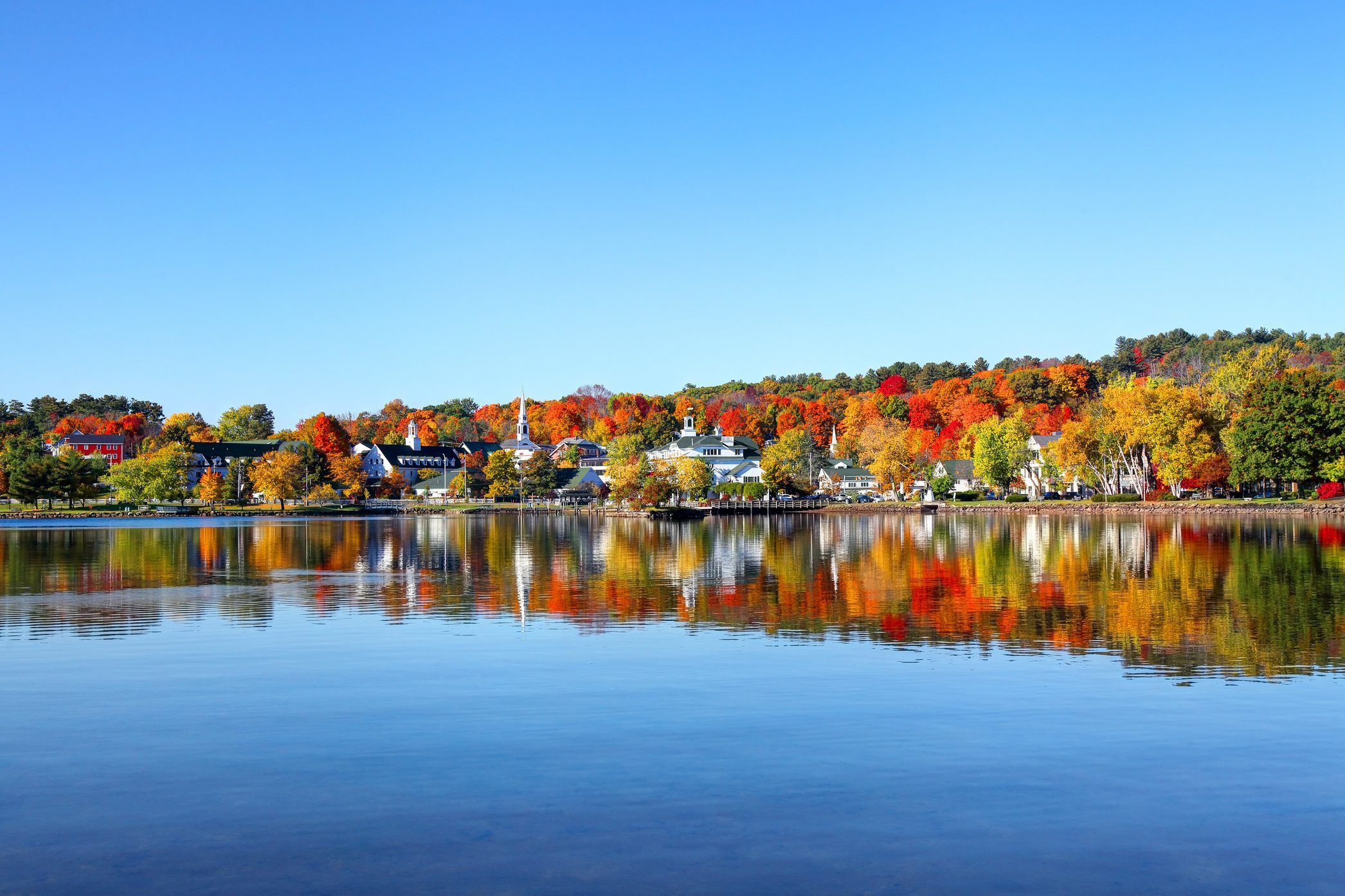 Trees display vibrant fall colors, reflected in a calm lake, with quaint buildings in the background under a clear blue sky.