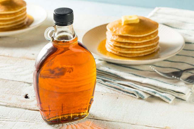 Bottle of maple syrup standing on table with pancakes in background, topped with butter on a white plate, on striped napkin.
