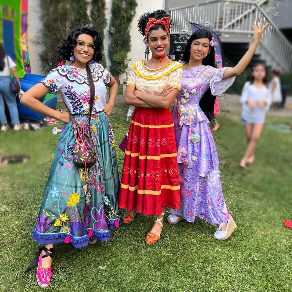 Three women pose in colorful, embroidered dresses, smiling on a grassy lawn with blurred figures and a staircase in the background.