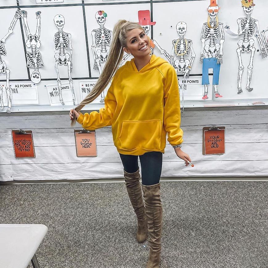 Person in a yellow hoodie playfully holds hair in a classroom with skeleton diagrams, motivational signs, and carpeted floor.