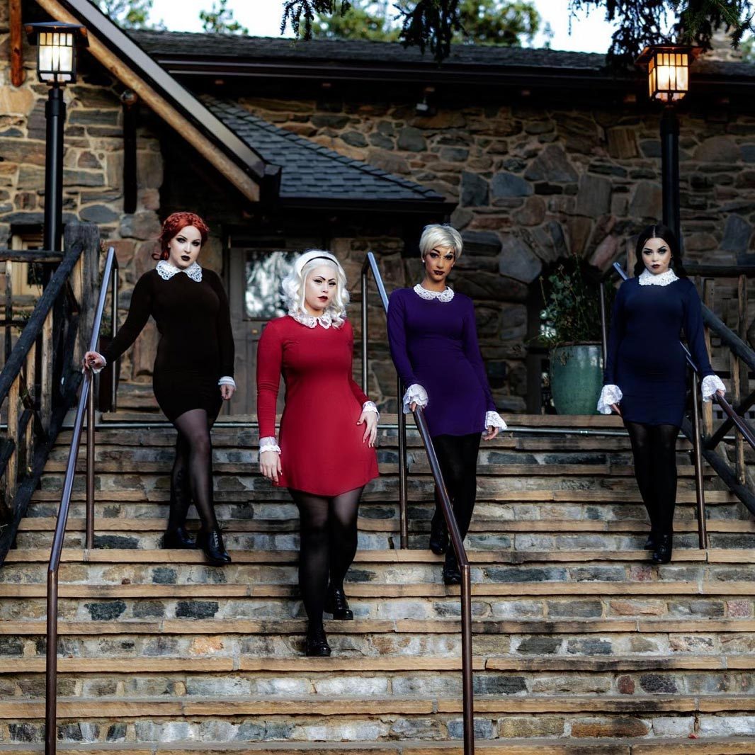 Four women in vintage-style dresses descend stone stairs, each wearing a different colored dress. A rustic stone building is in the background.