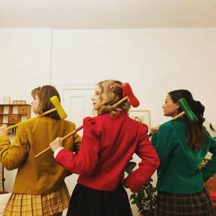 Three people stand with croquet mallets over shoulders, wearing red, yellow, and green jackets in a room with white walls and plants.