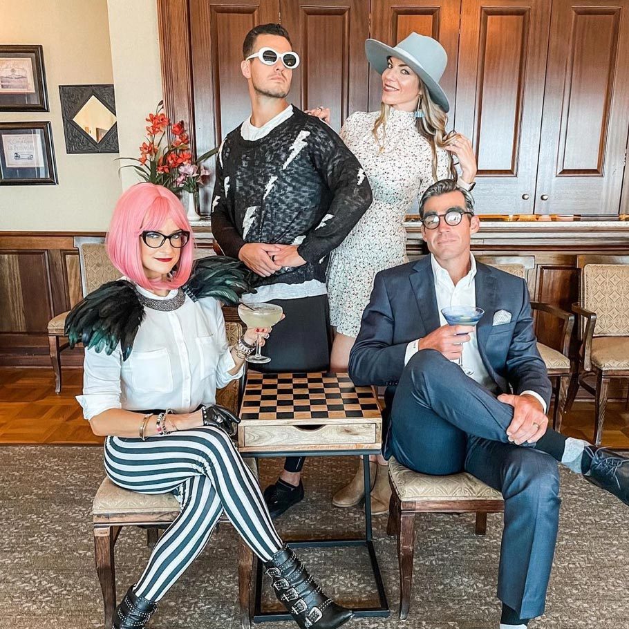 Four people with eclectic clothing pose around a chess table, holding drinks, in a room with wooden cabinets and framed pictures.