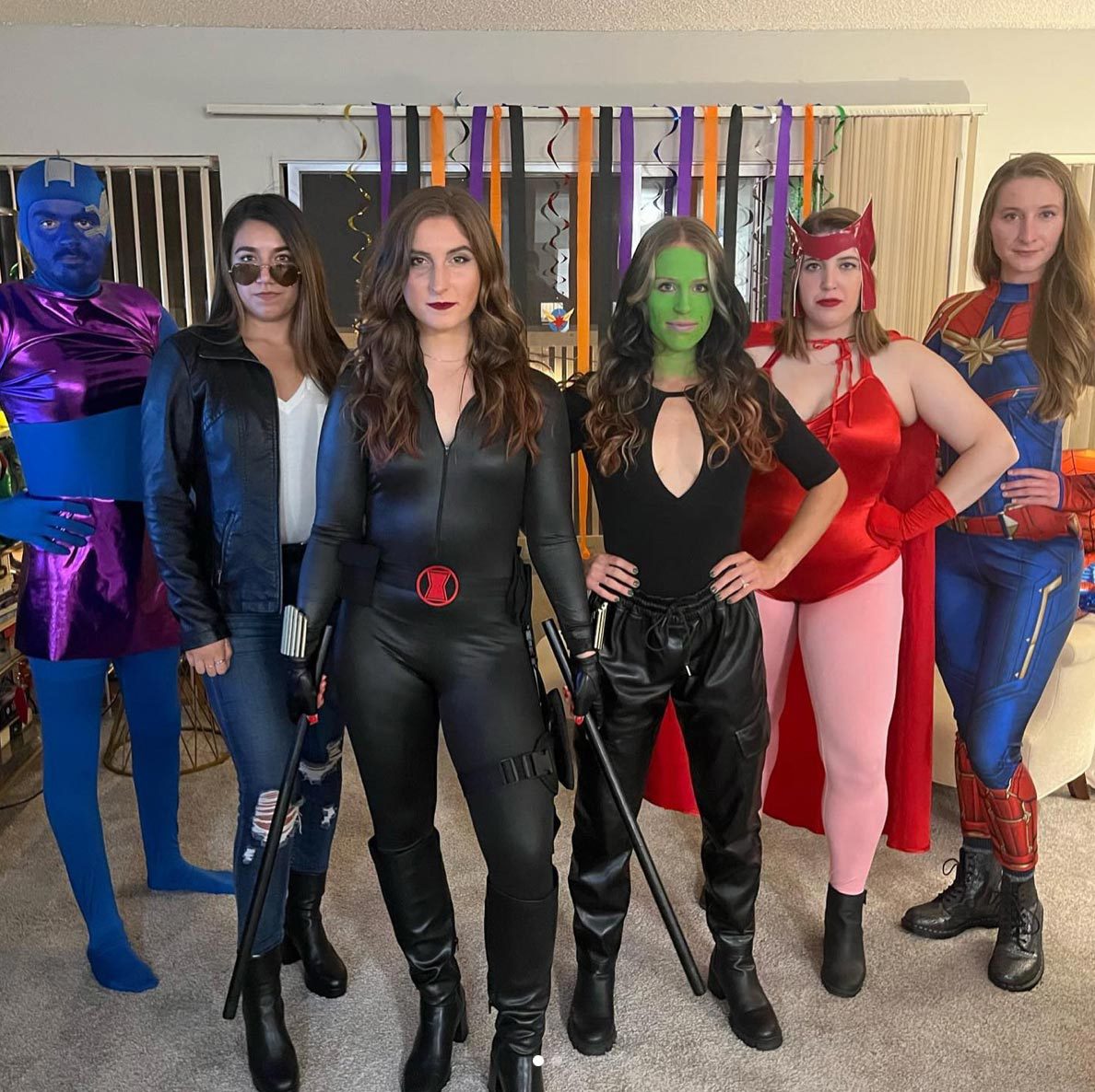 Five people wear superhero costumes, posing confidently in a living room decorated with colorful hanging ribbons.