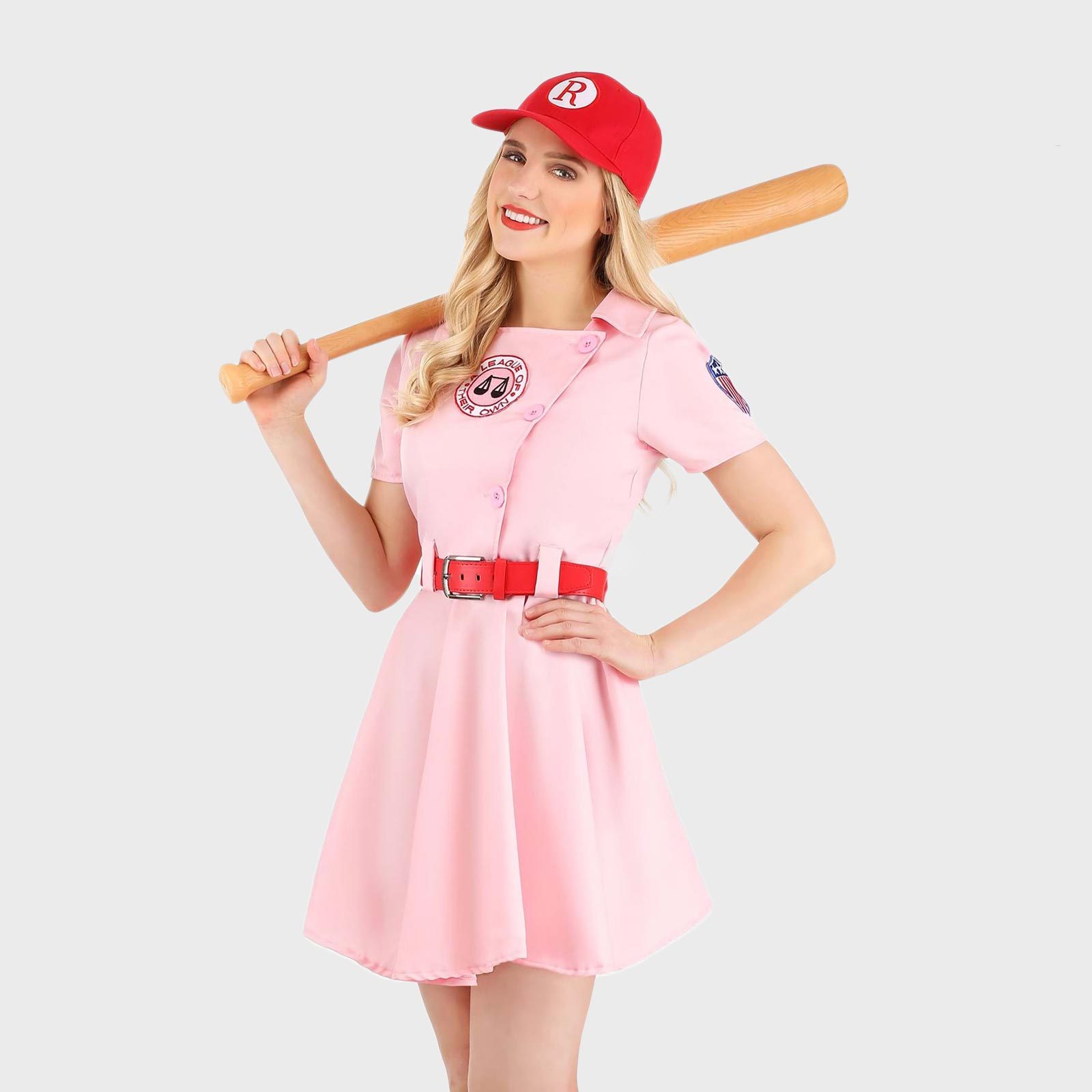 A person holds a baseball bat over the shoulder, wearing a pink dress and red cap in a neutral, brightly lit space.