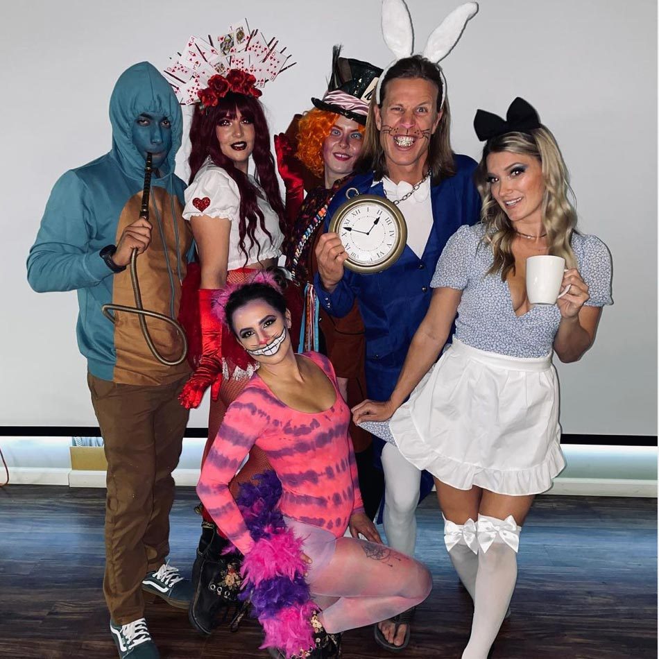 Six people wear whimsical costumes, posing indoors. One person holds a clock, another a cup, and several have colorful, fantastical outfits and accessories.
