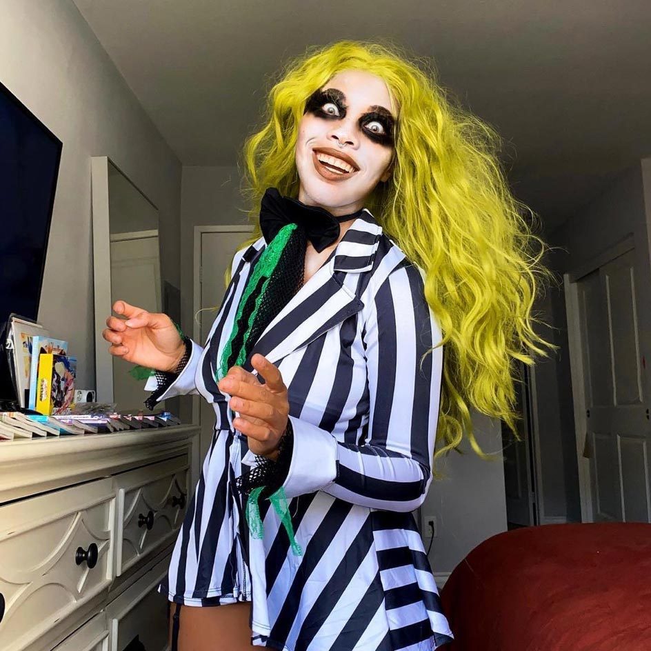 Individual dressed in a black-and-white striped costume with vibrant yellow hair, posing in a room with a dresser and television.