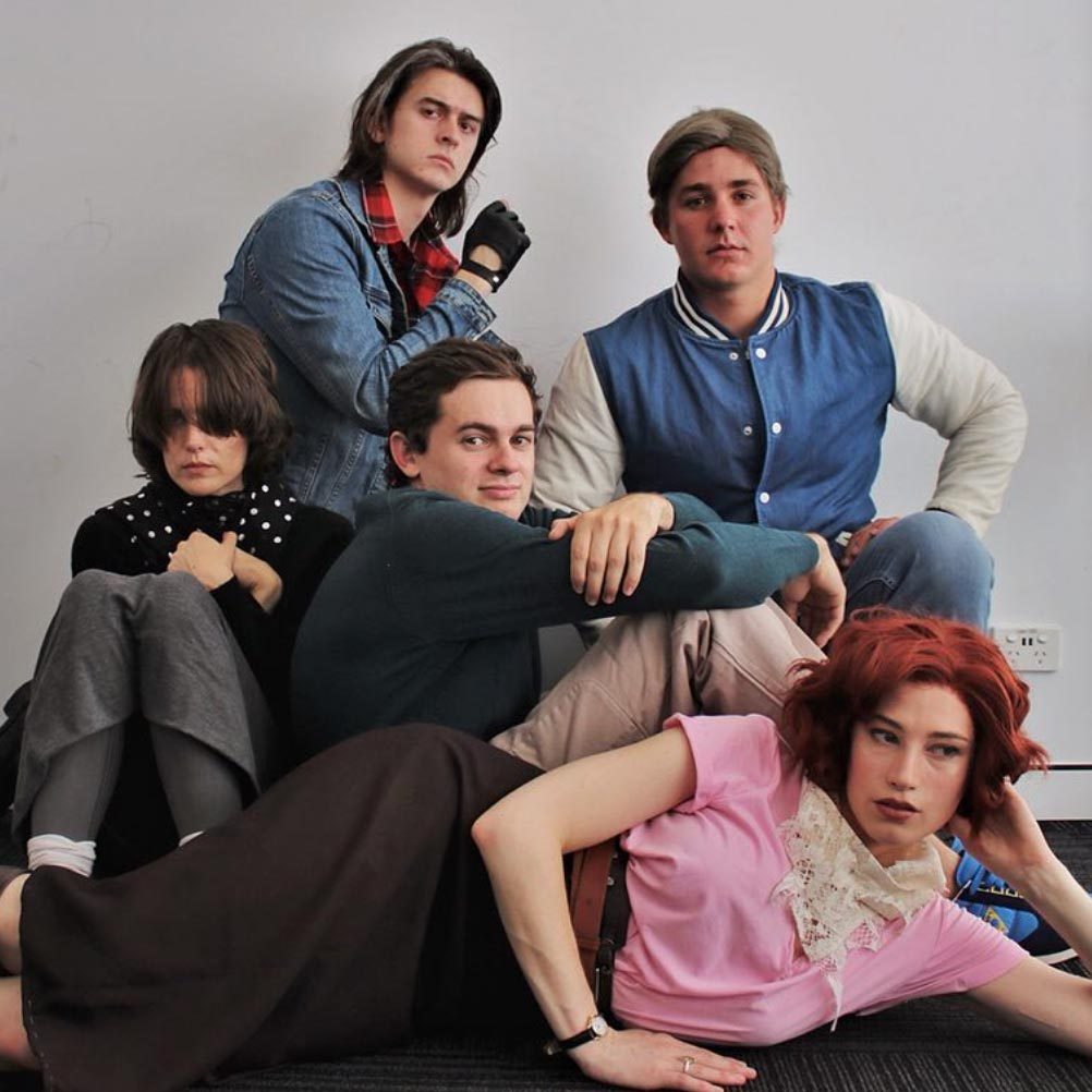 Five people pose sitting and leaning against each other, wearing casual and retro-themed clothes, in a room with a plain white wall and dark flooring.