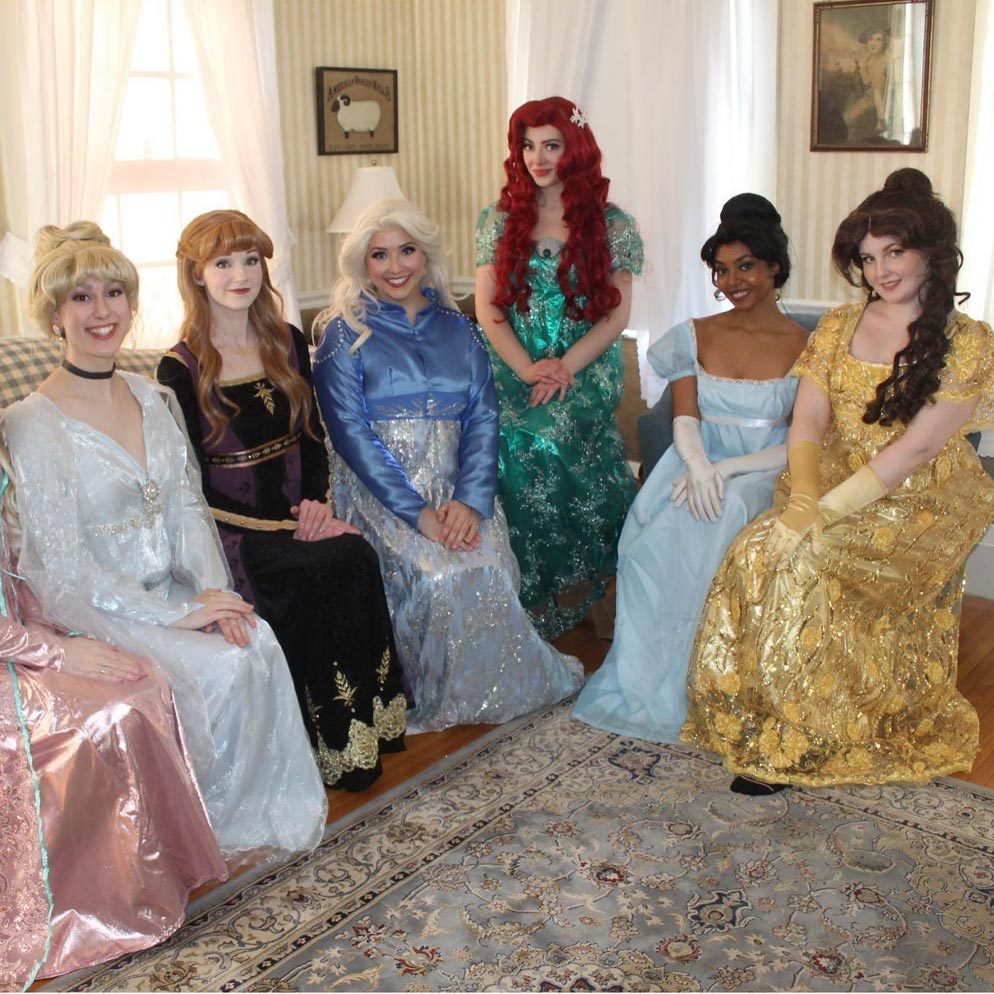 Women dressed as princesses sit smiling in a cozy, wallpapered room with a decorative rug and framed artwork on the walls.
